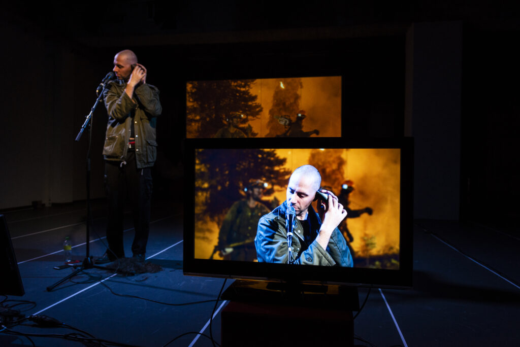 Szenenfoto der Premiere. Im Vordergund ein Bildschirm, auf dem das Kamerabild dessen zu sehen ist, was wir hintert dem Bildschirm sehen: Vor einer Projektion brennender Bäume rasiert der Performer seinen Kopf.
