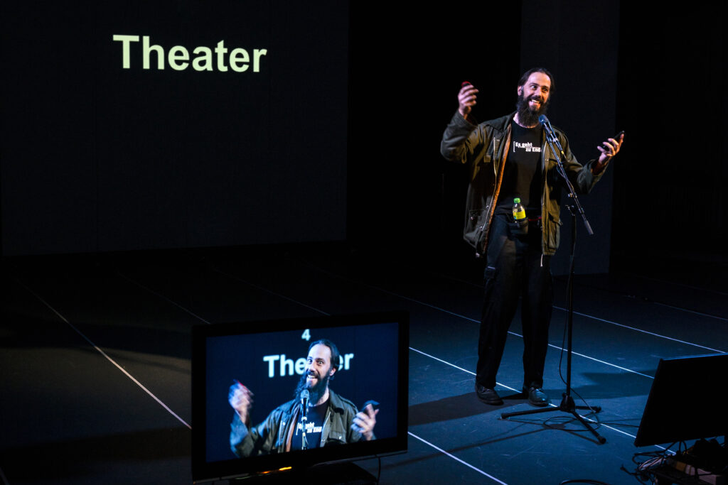 Szenenfoto. Auf einer Projektion steht das Wort "Theater", davor gestikulierend/grimmassierend der Performer. Ein Bildschirm im Vordergrund zeigt das Kamerabild dieser Szene.