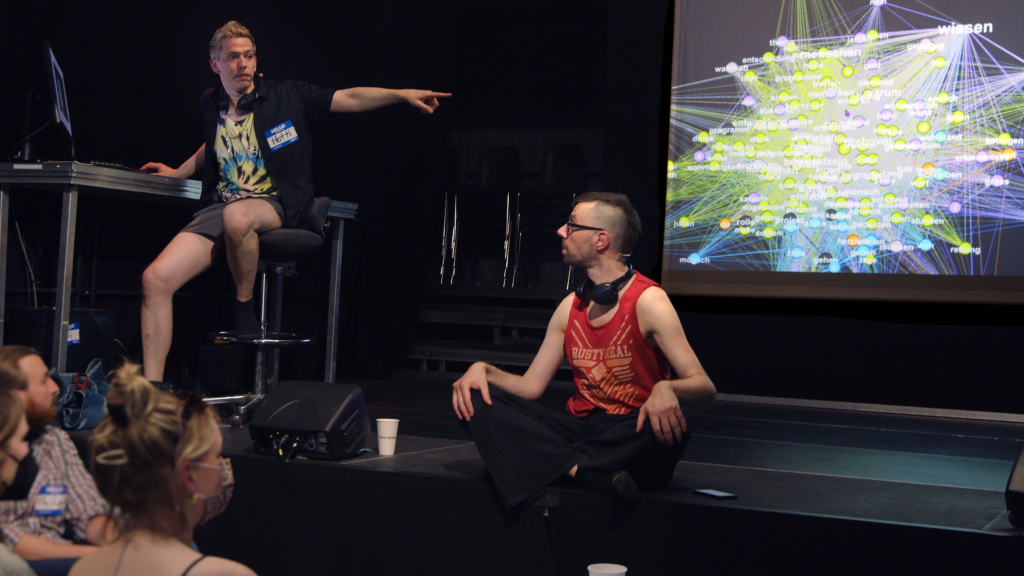 Hotti, links auf einem hohen Hocker sitzen, zeigt mit dem linken gestreckten Arm auf eine bunte Netzwerkvisualisierung auf einer Projektionsleinwand im Bildhintergrund. Arne sitzt davor und schaut zu ihm. Links vorne am Bildrand sehen wir Köpfe des Publikums, ihre Gesichter sind nicht identifizierbar.