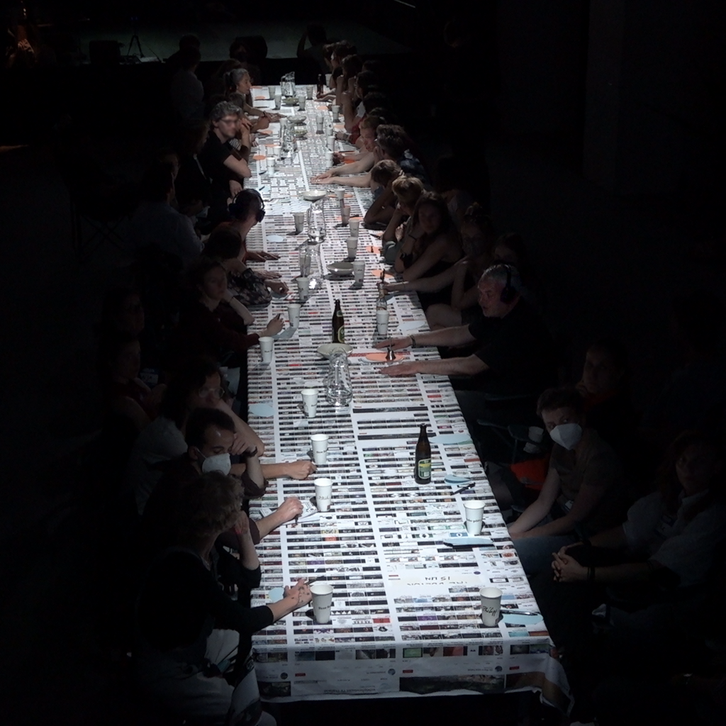 In einem dunklen Raum eine hell beleuchtete lange Tafel, an der ca. 30 Menschen sitzen. DIe Tischfläche ist komplett mit einer langen Stoffbahn bedeckt, auf die in vier Spalten die Inhalte mehrerer YouTuibe-Kanäle mit tausenden Videothumbnails gedruckt sind. Darauf stehen Flaschen und Becher.