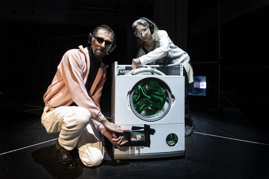 Arne und Marina an einer präparierten und innen LED-grün leuchtenden Waschmaschine, es sieht etwas albern aus.