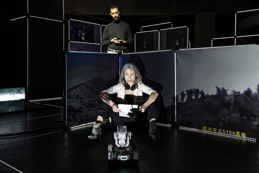 Marina sitzt vor hüfthohen Projektionswänden und spricht über chinesischen Cyberwar, von vorne filmt sie ein fahrbares Roboter-Auto. Hinter ihr steht Arne und schaut/hört zu. In der Tiefe staffelb sich weitere Projektionswände unterschiedlicher Höhe und Breite.